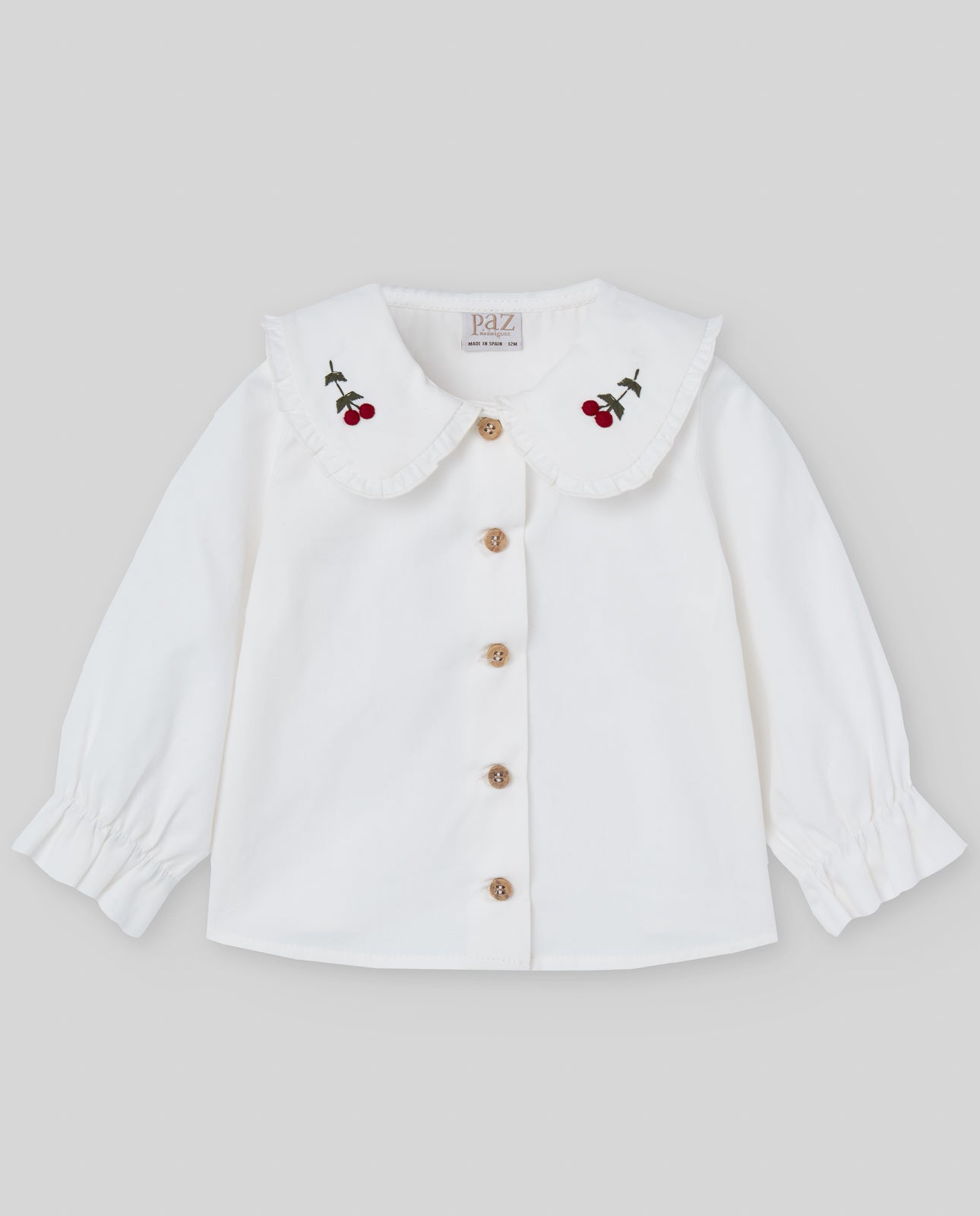 White blouse with cherry embroidery and gold buttons on a light gray background