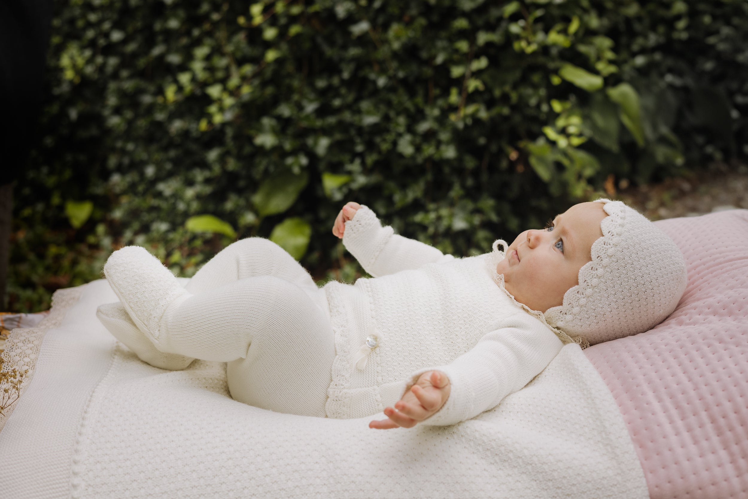 Baby lying on a blanket with a pink blanket and greenery in the background