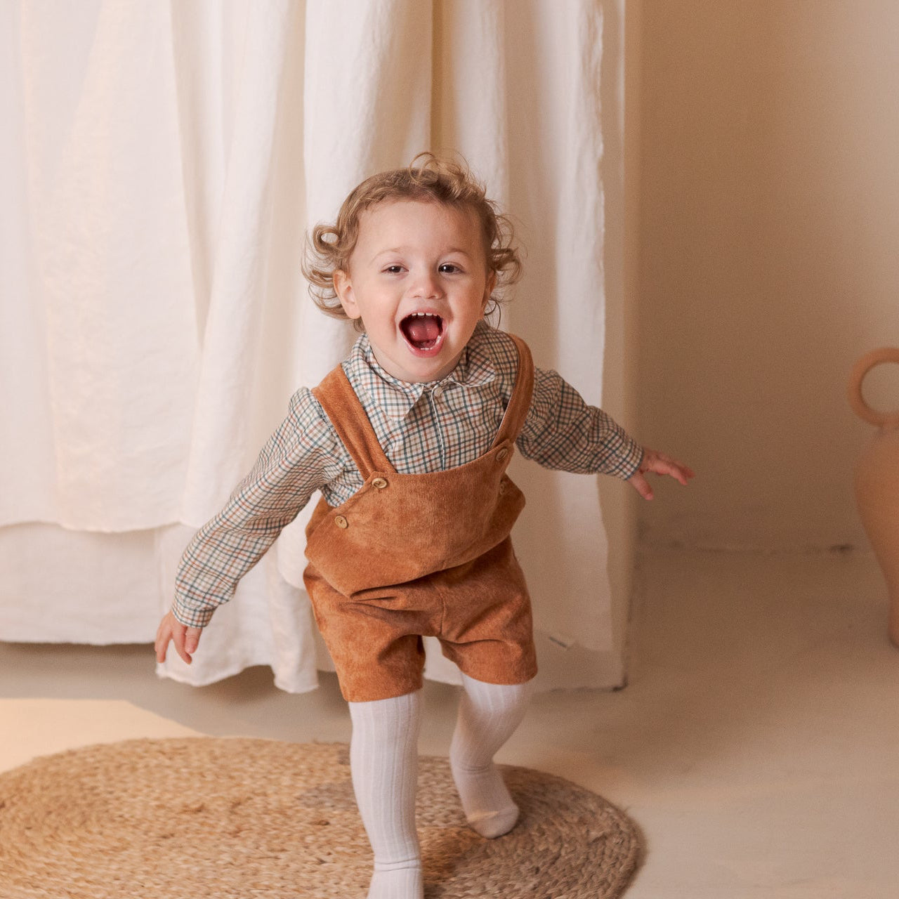 Child in a brown outfit standing in a room with a vase and curtain