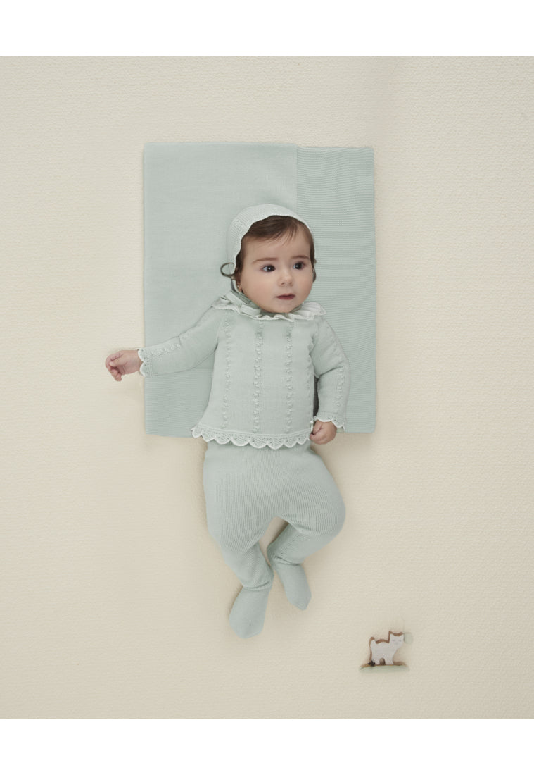 Baby in a light green outfit standing on a white surface with a light blue blanket behind.