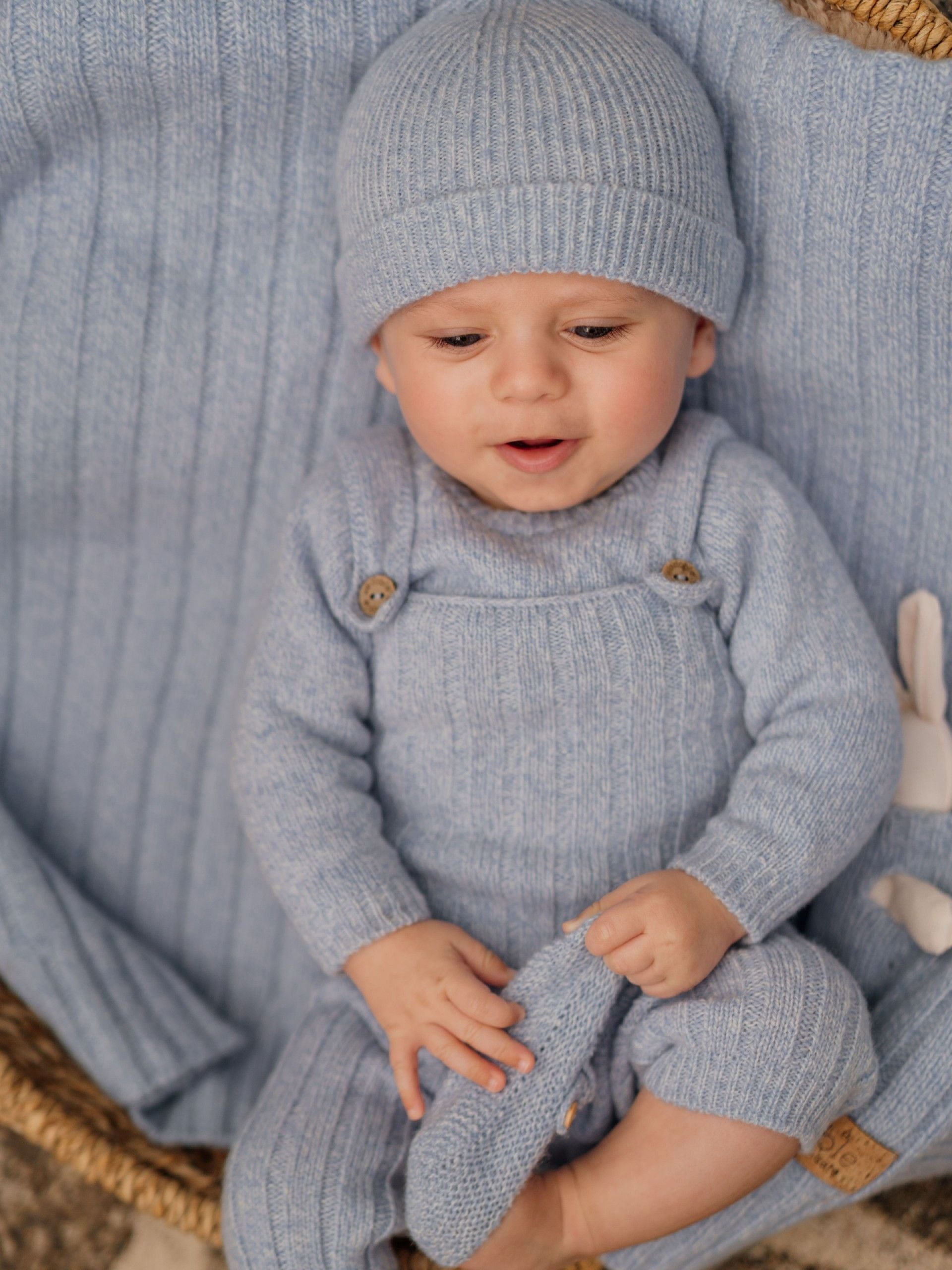 Baby in a matching knit outfit with a light blue hat and romper, sitting on a textured surface.
