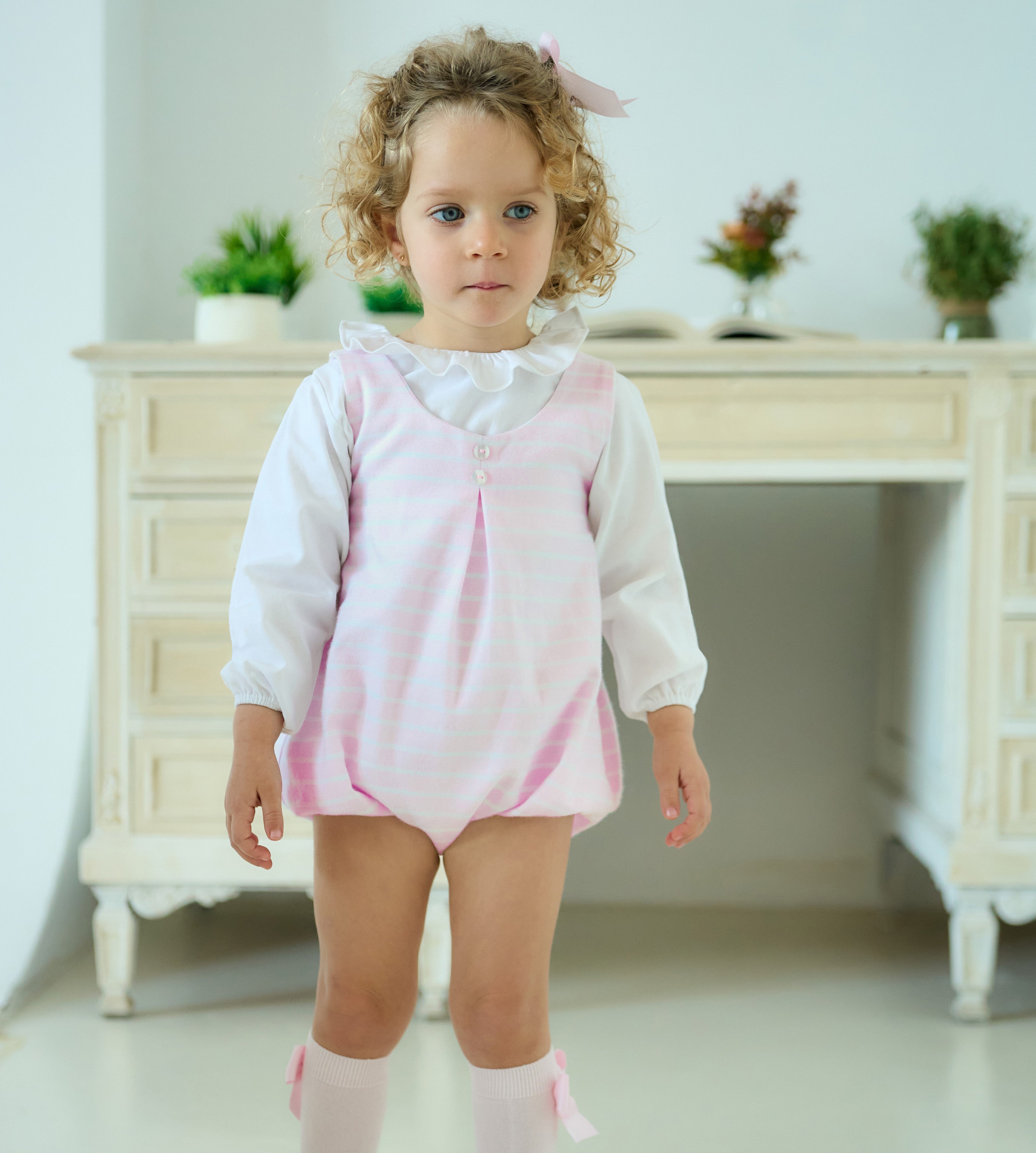 Young girl wearing a pink and white outfit standing in a bright room with white furniture and plants.