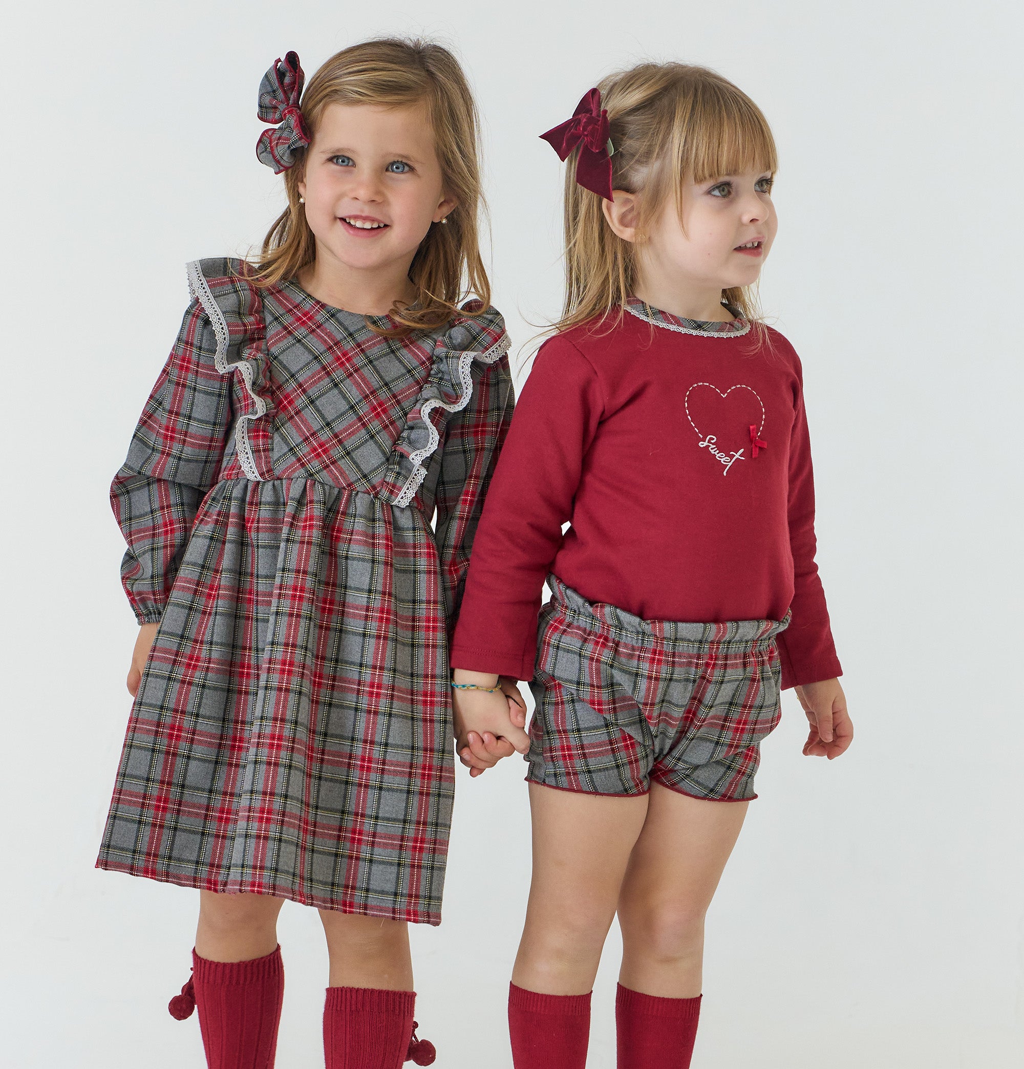 Two young girls holding hands, one in a plaid dress and the other in a red top and plaid shorts, on a white background.