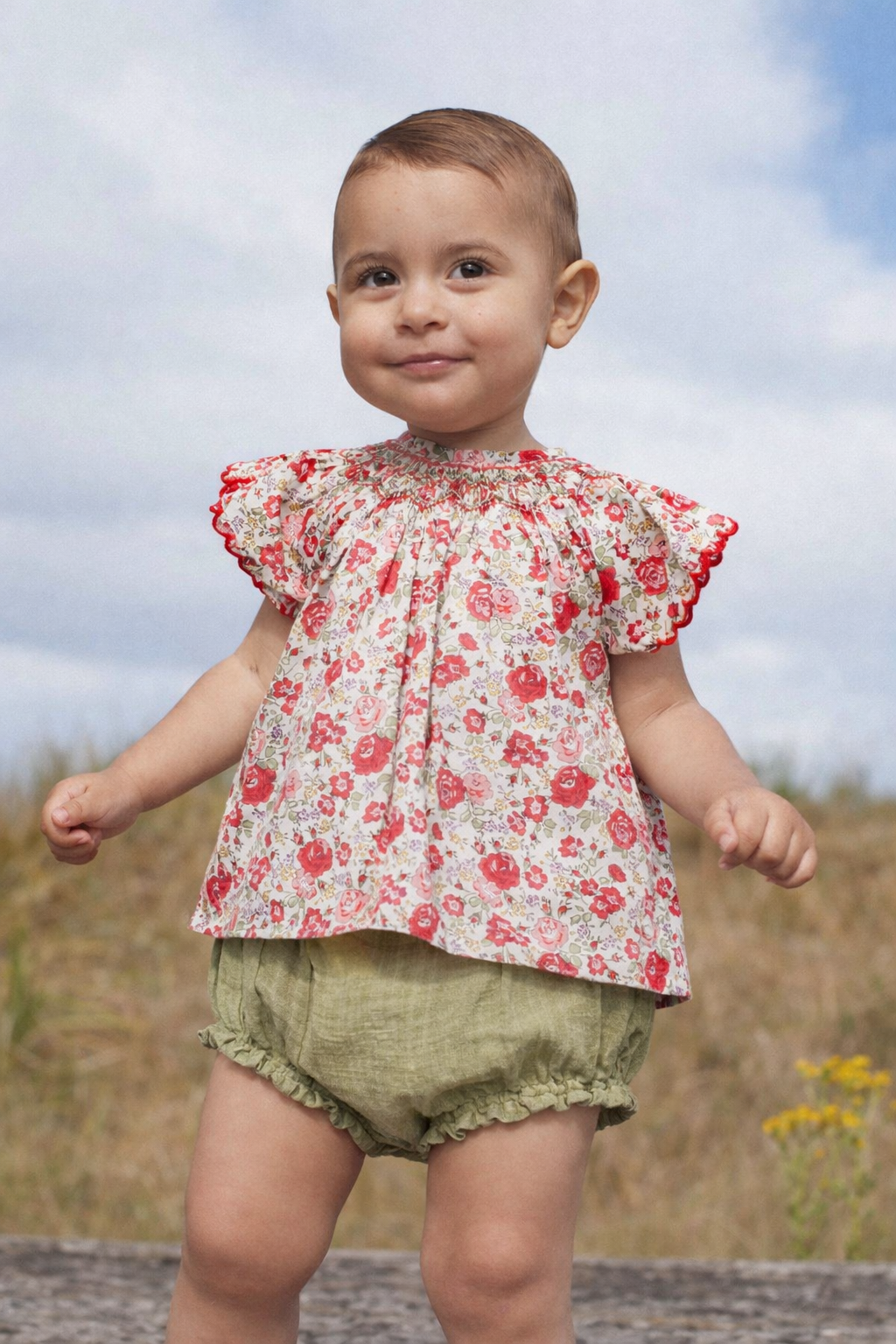 Child wearing a floral top and green bloomers outdoors with a blue sky background