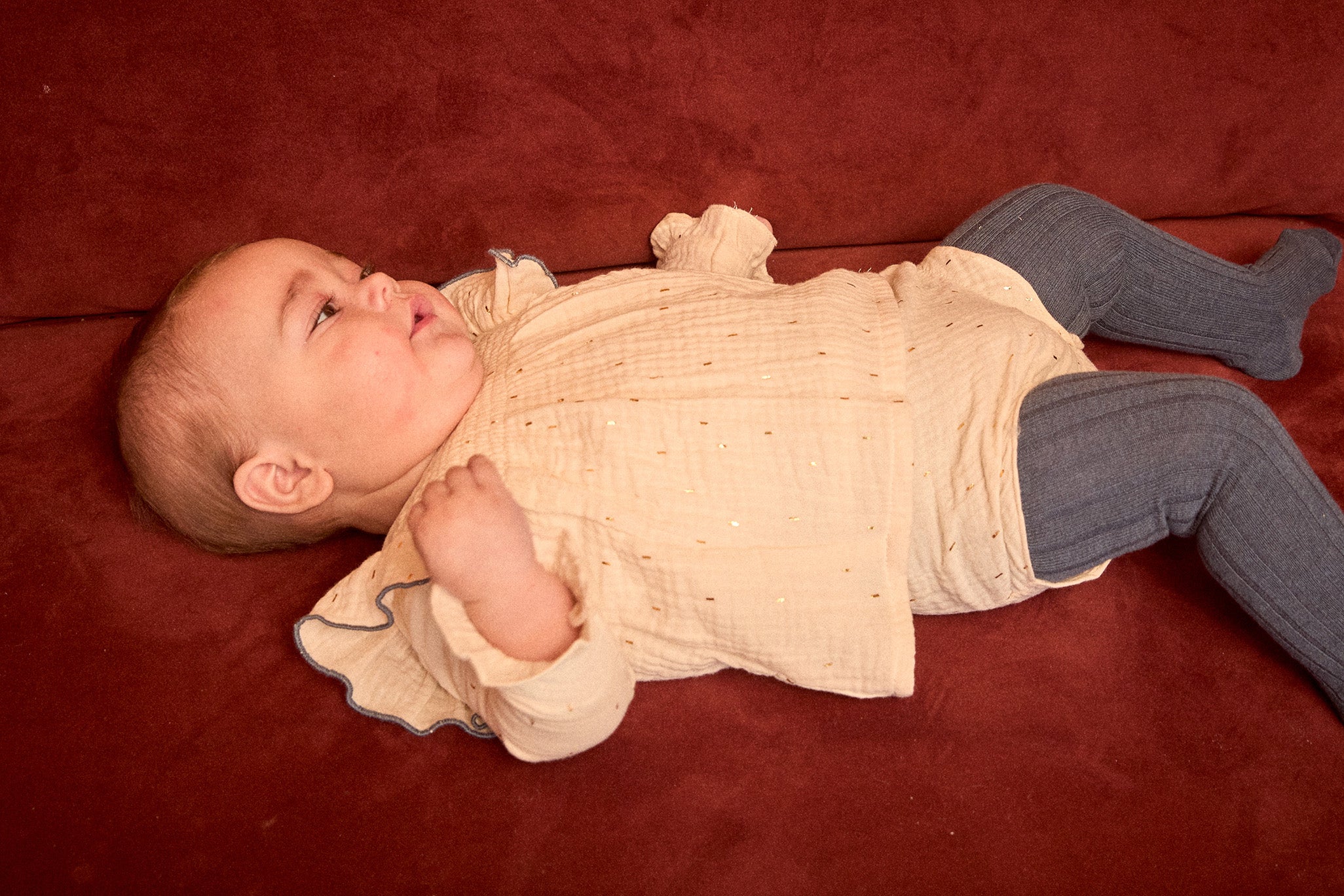 Baby lying on a red blanket wearing a beige outfit and gray pants.