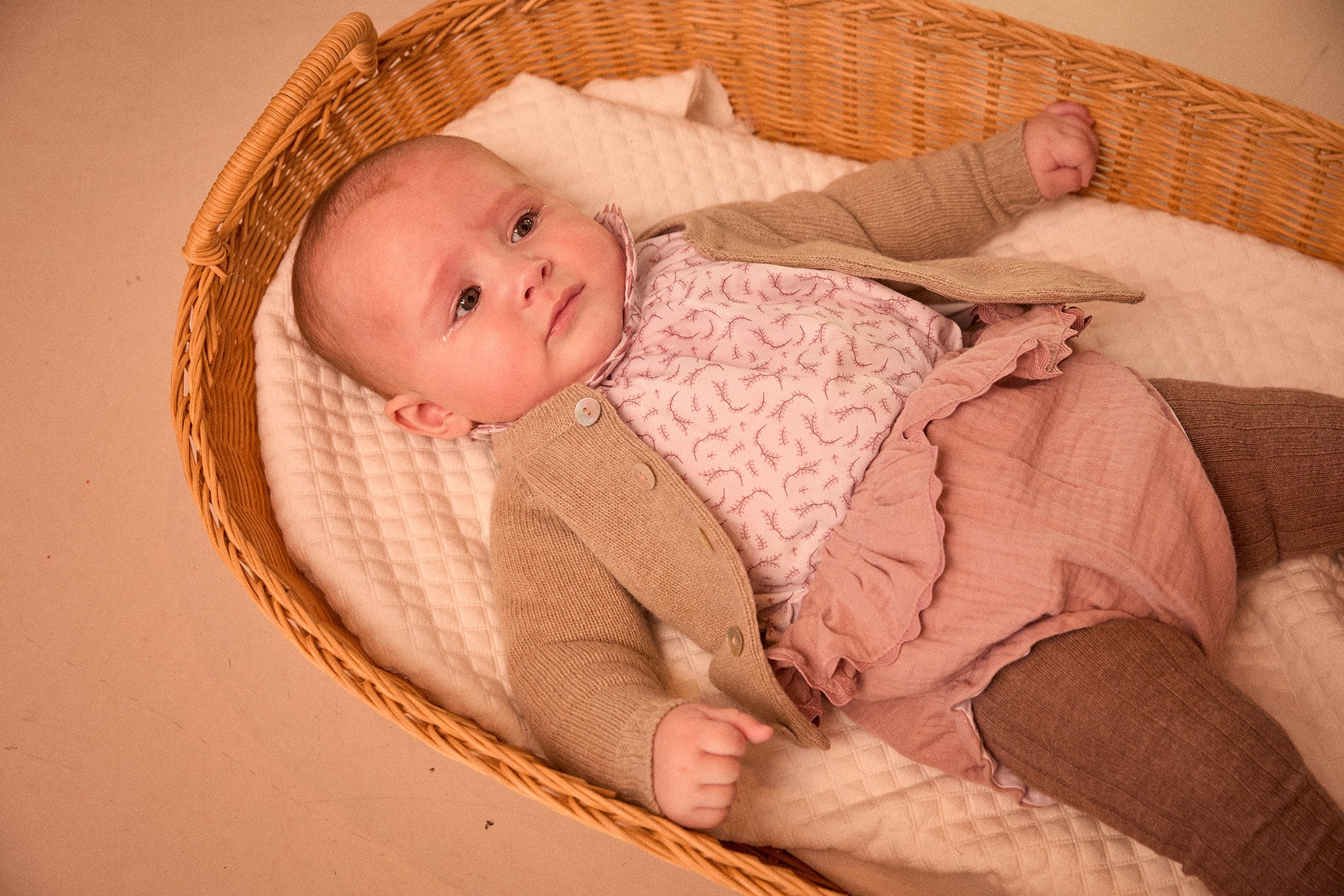 Baby in a wicker crib wearing a brown coat and pink dress.
