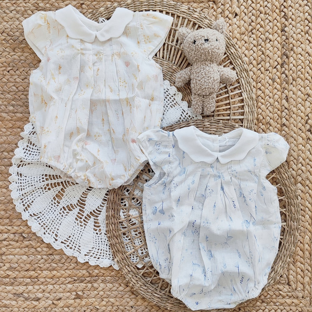 Two baby rompers with floral patterns on a woven surface with a teddy bear.