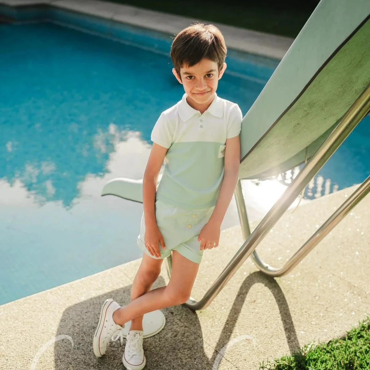 Child standing by a pool with a green lawn and pool deck in the background