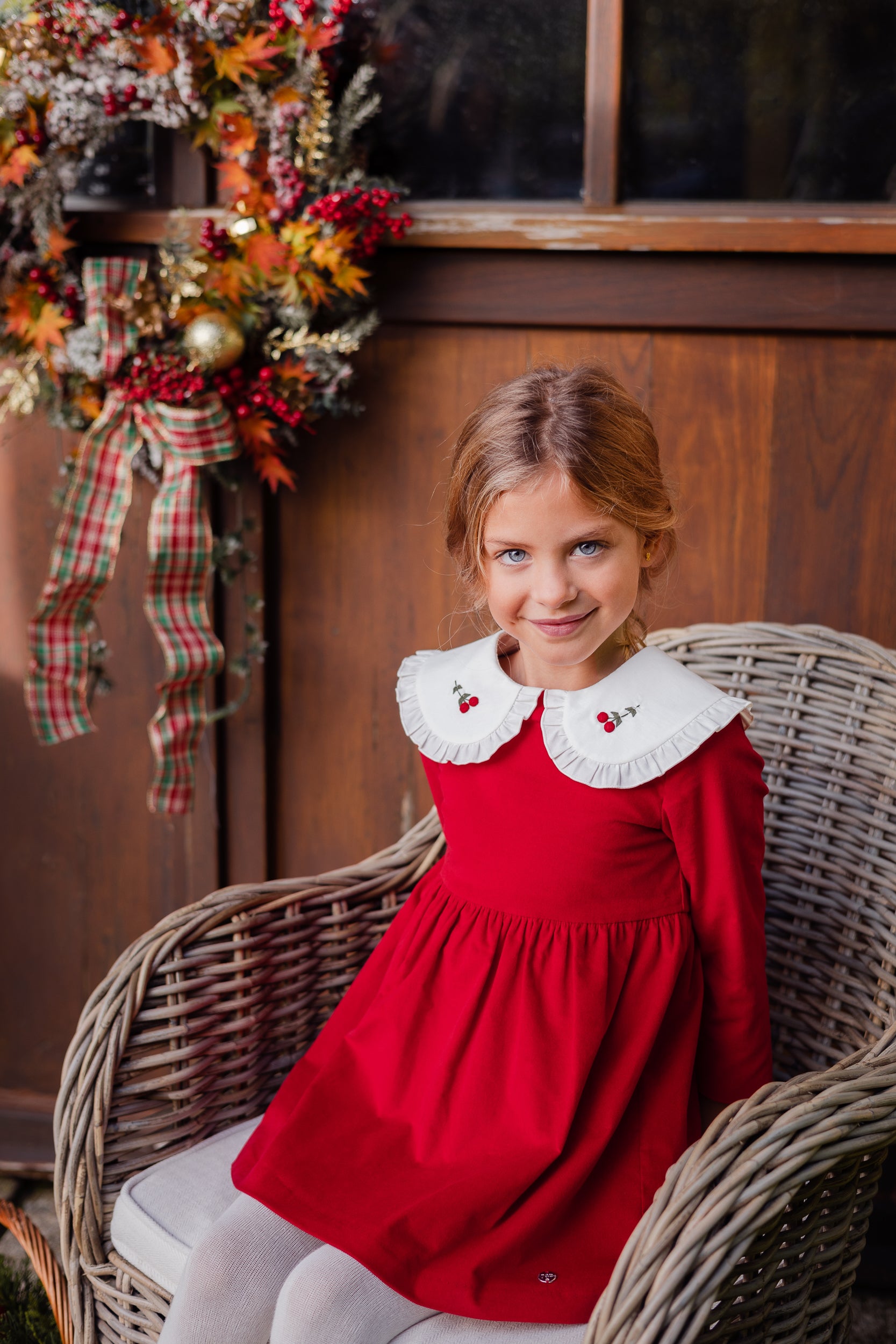 Young girl in a red dress with white collar sitting on a wicker chair in front of a wooden wall with decorative elements.