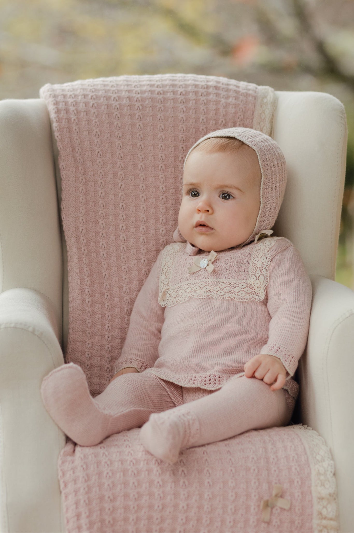 Baby in pink outfit sitting on a white chair with a pink blanket in an outdoor setting