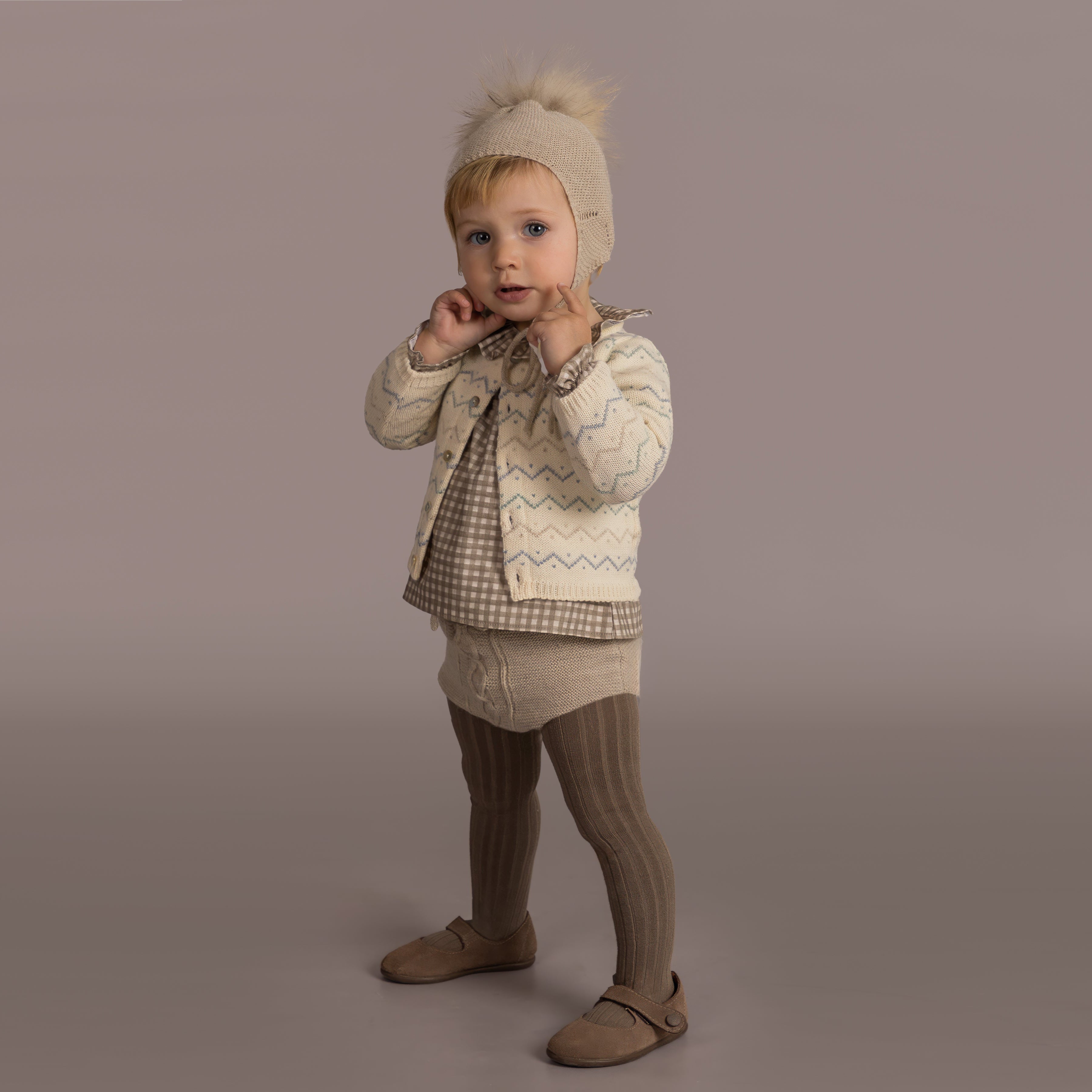 Child wearing a beige knit outfit with a matching hat on a plain background