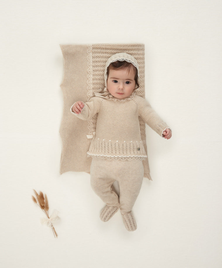 Baby in a beige knitted outfit lying on a white blanket with a textured pillow.
