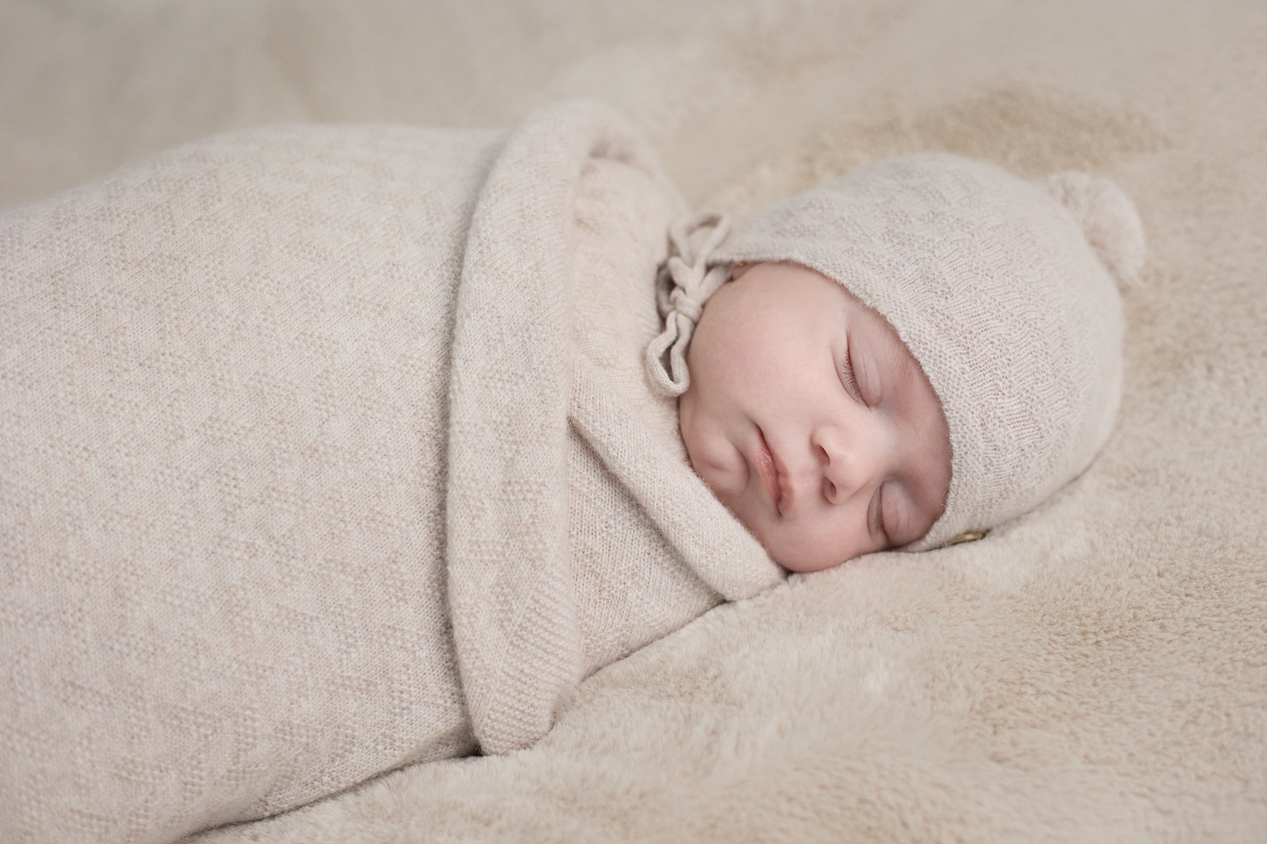 Newborn baby wrapped in a beige blanket and bonnet on a soft surface