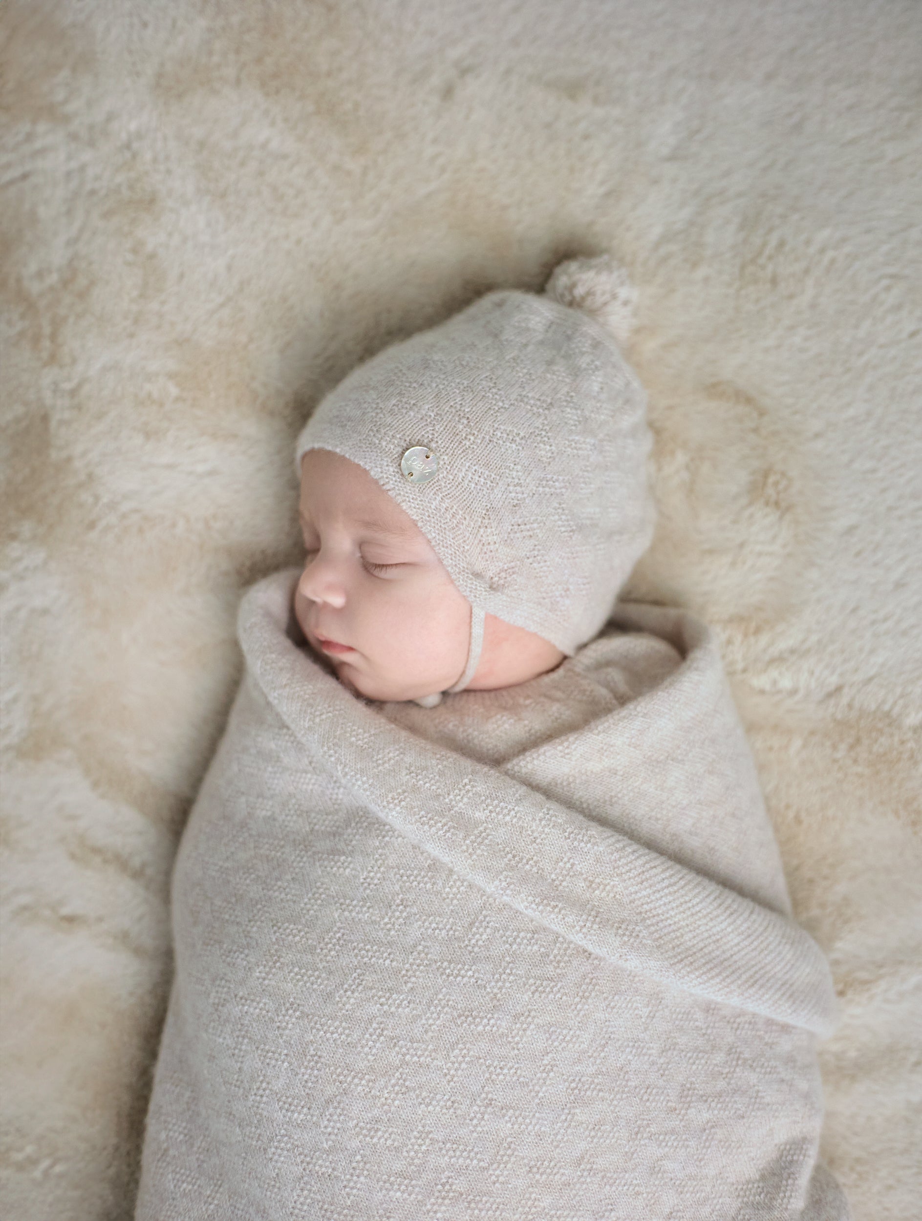 Newborn baby swaddled in a light gray blanket with a matching hat on a soft beige surface.
