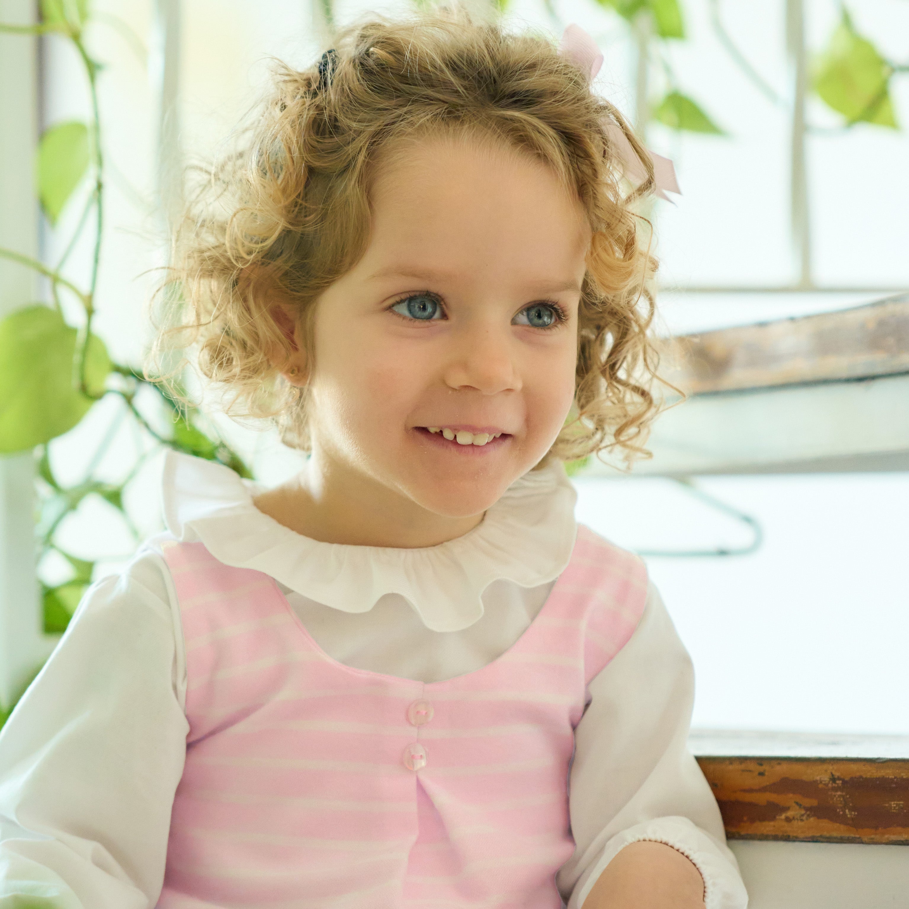 Young girl with curly hair wearing a pink dress with a white collar, sitting indoors.