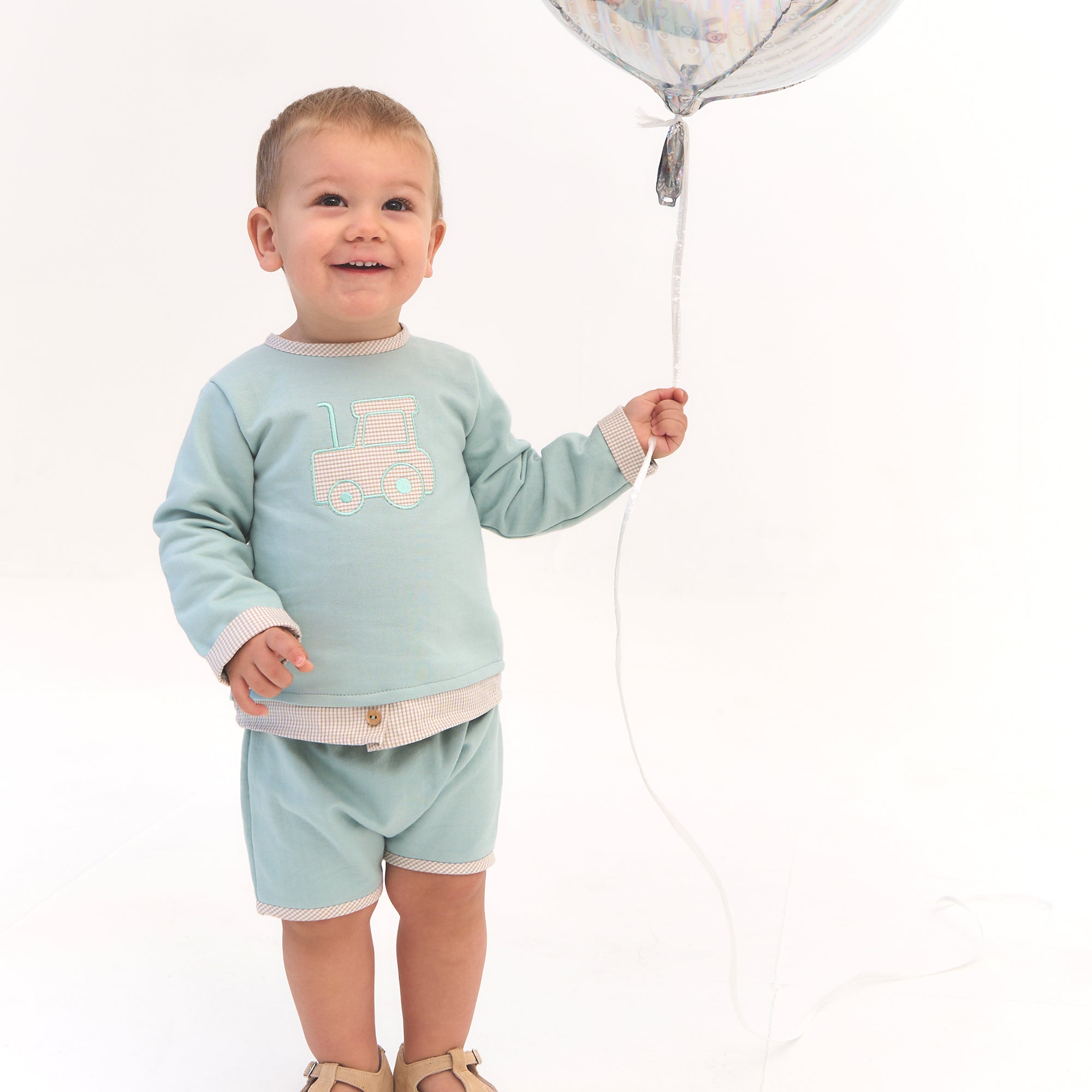 Child wearing a light blue outfit with a train design, holding a balloon against a white background