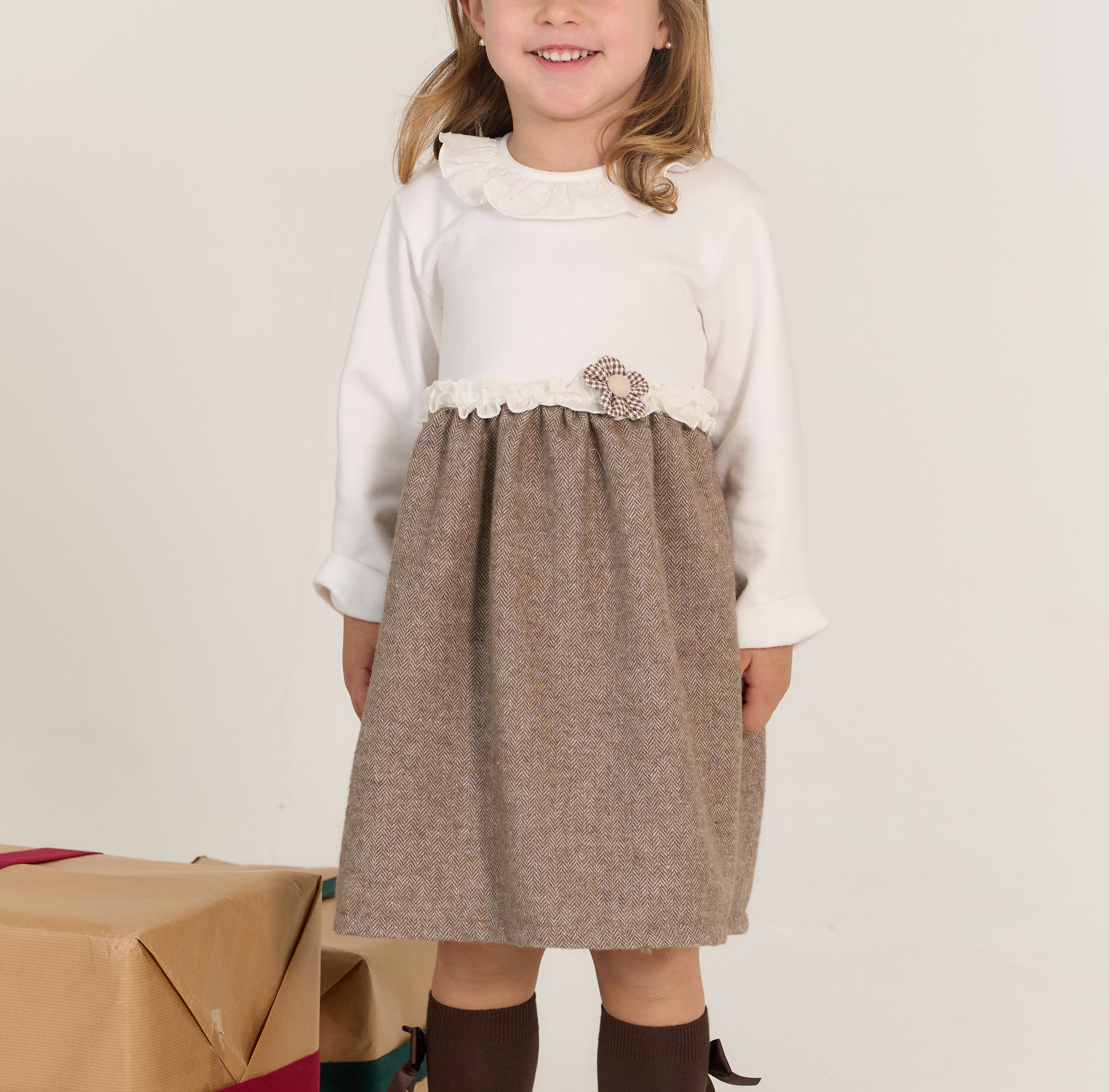 Young girl wearing a white long-sleeve top and brown dress with a decorative brooch, standing against a plain background.