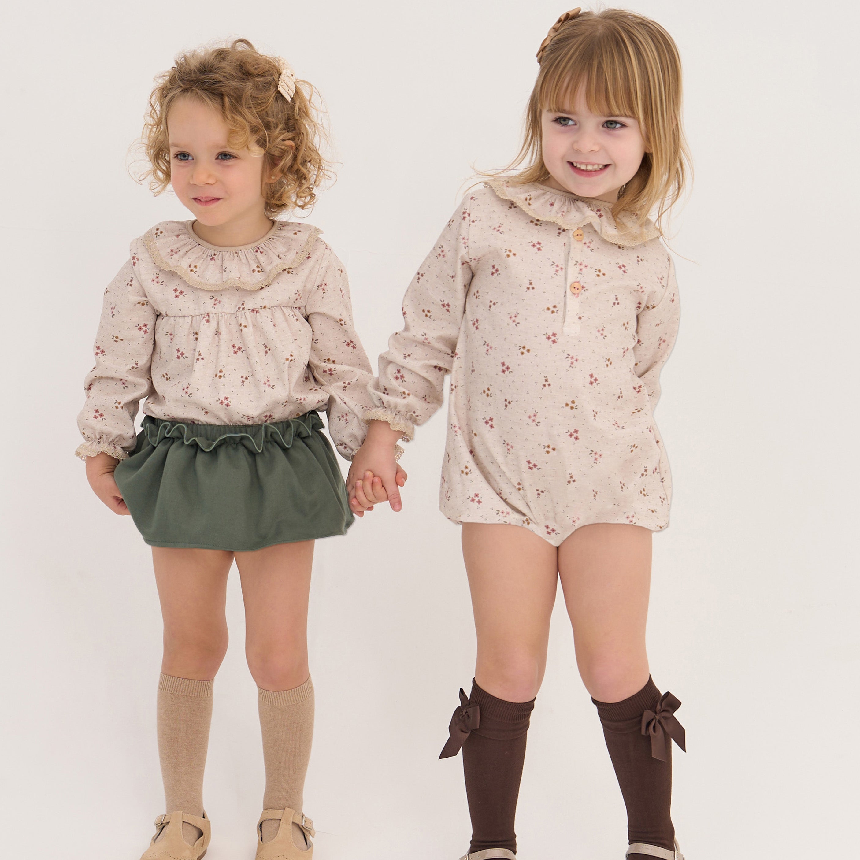 Two young girls holding hands, wearing matching outfits with floral patterns on a plain background.