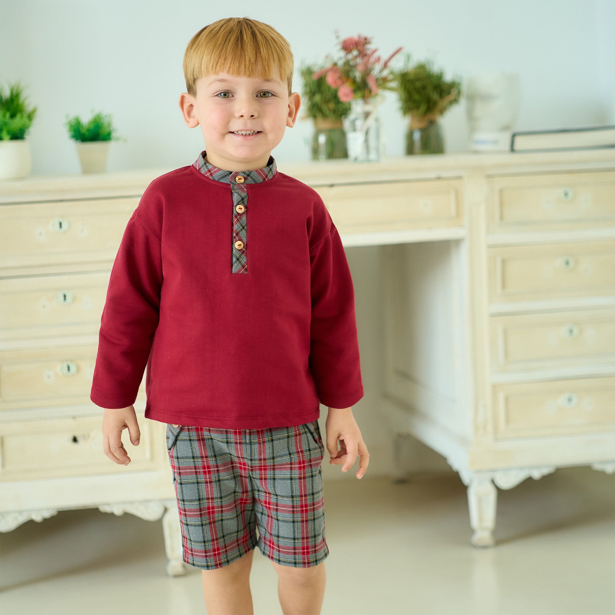 Child wearing a red sweater and plaid shorts standing in a room with a dresser and plants.