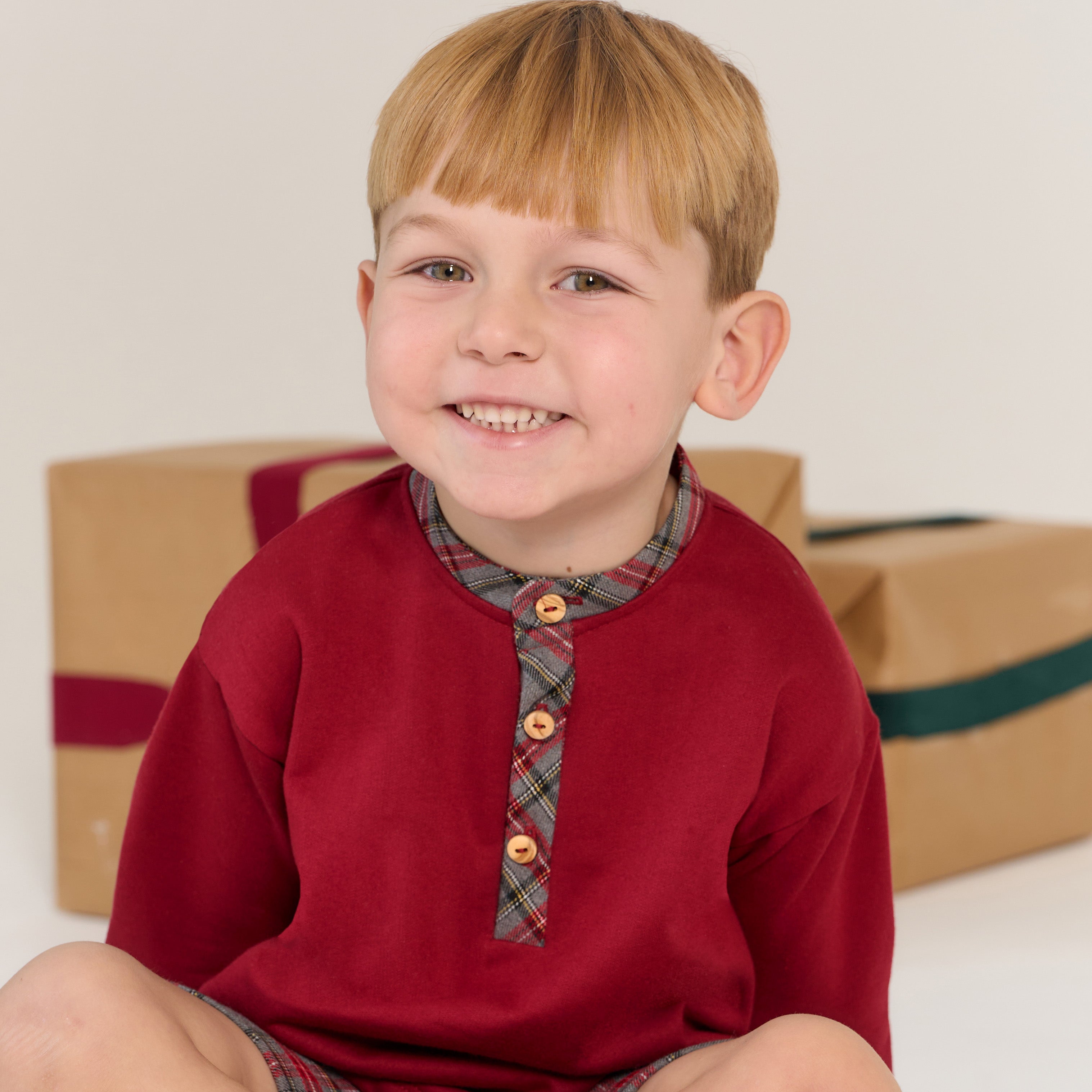 Young boy wearing a red shirt with plaid collar and buttons, sitting in front of gift boxes.