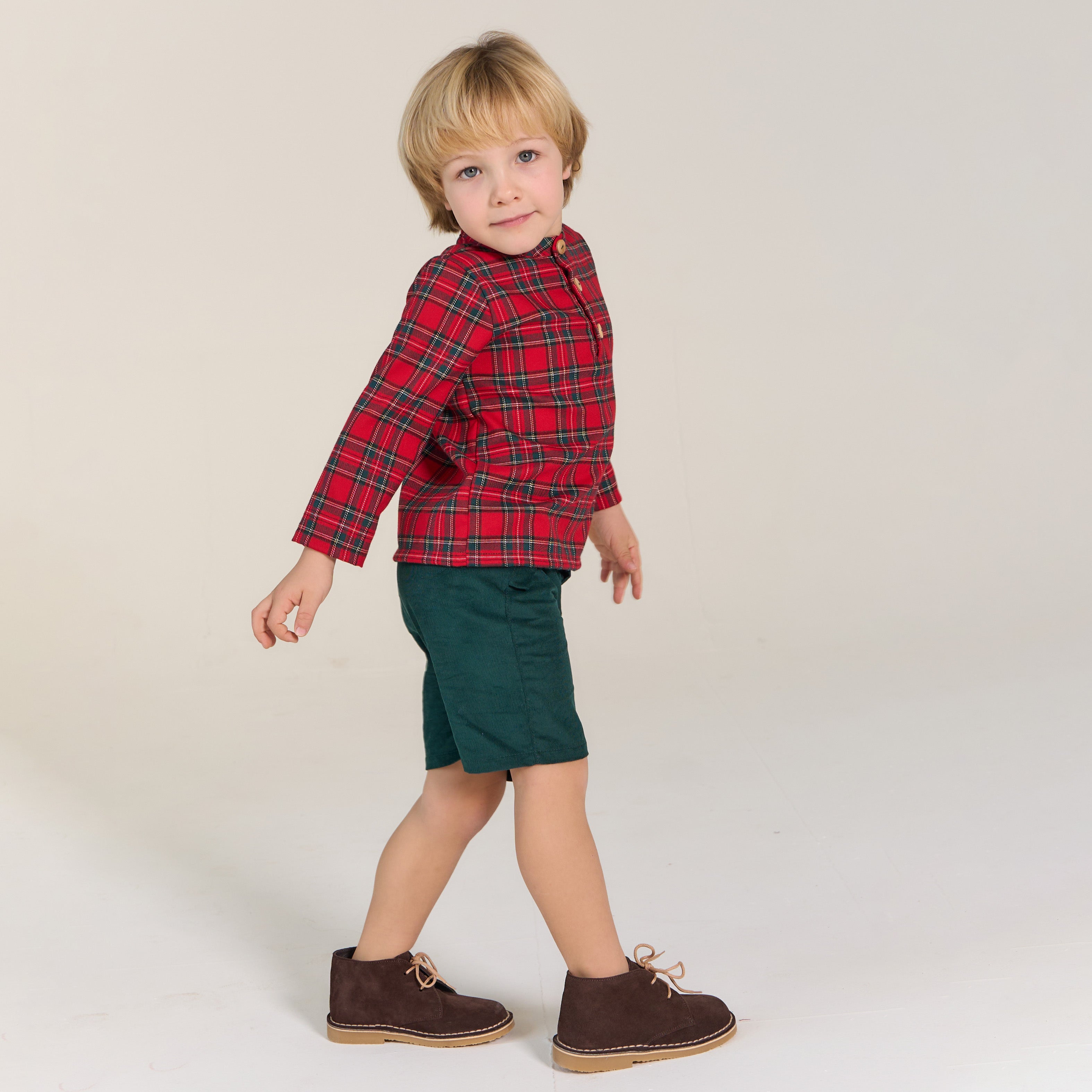 Child wearing a red plaid shirt, green shorts, and brown shoes on a plain background