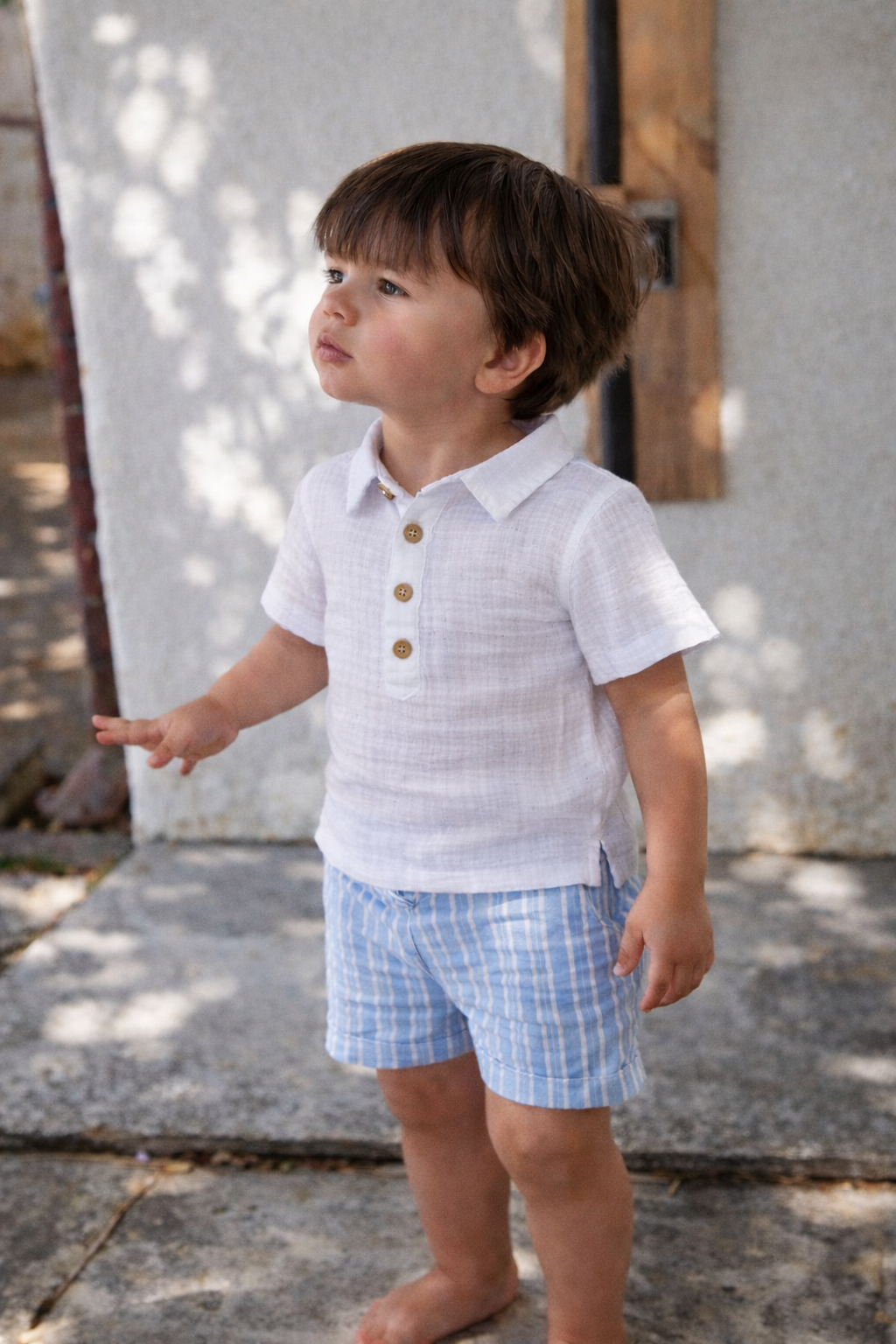 Child wearing a white shirt with wooden buttons and blue checkered shorts outdoors.