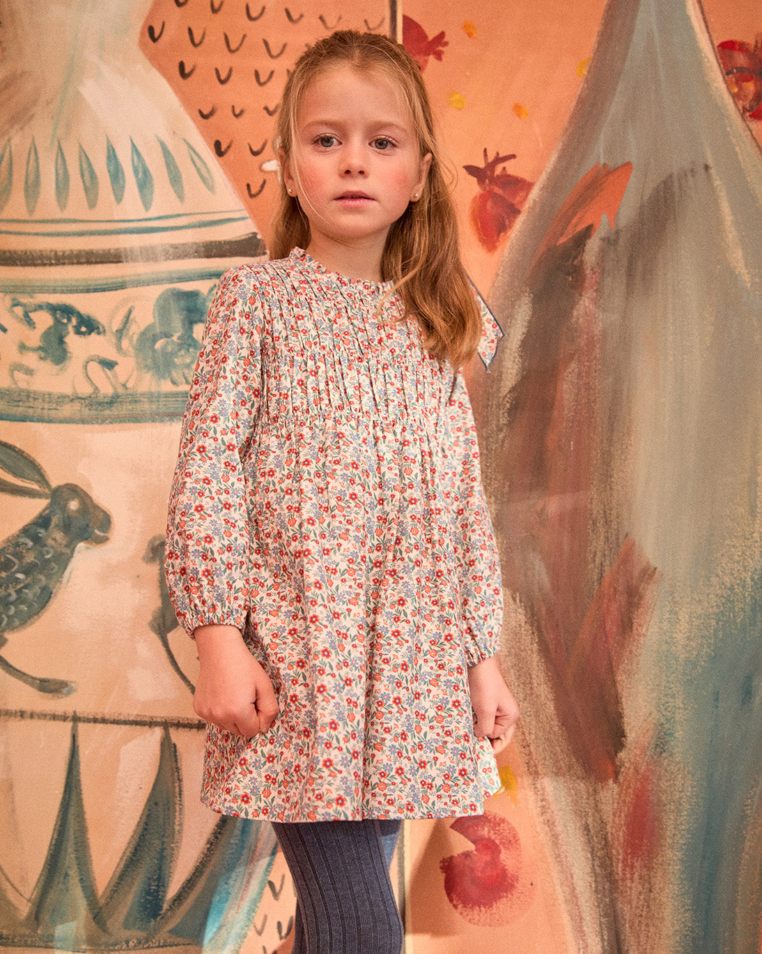 Young girl wearing a floral dress standing in front of a colorful wall with abstract designs.