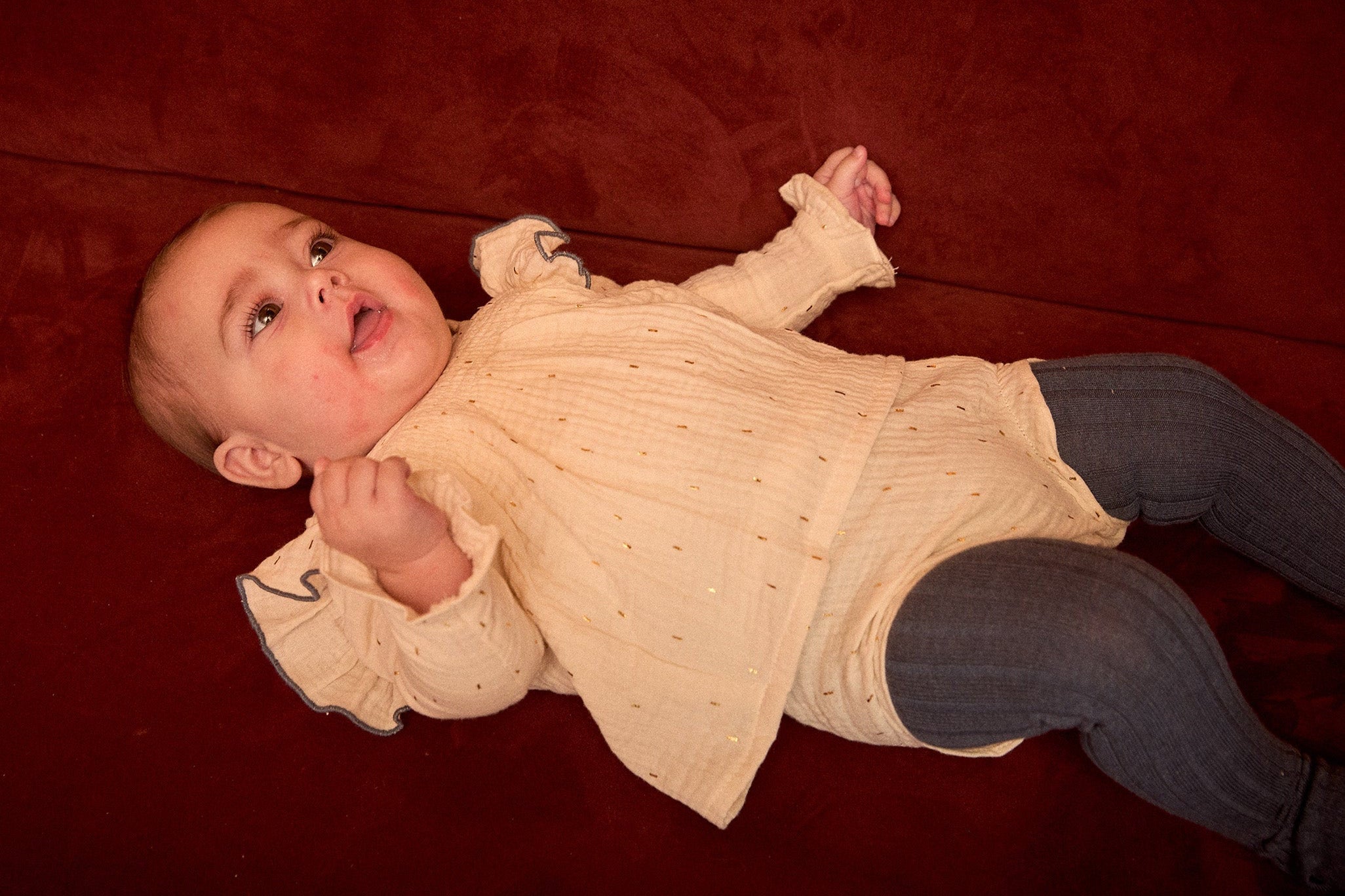 Baby lying on a red couch wearing a beige sweater and dark pants.