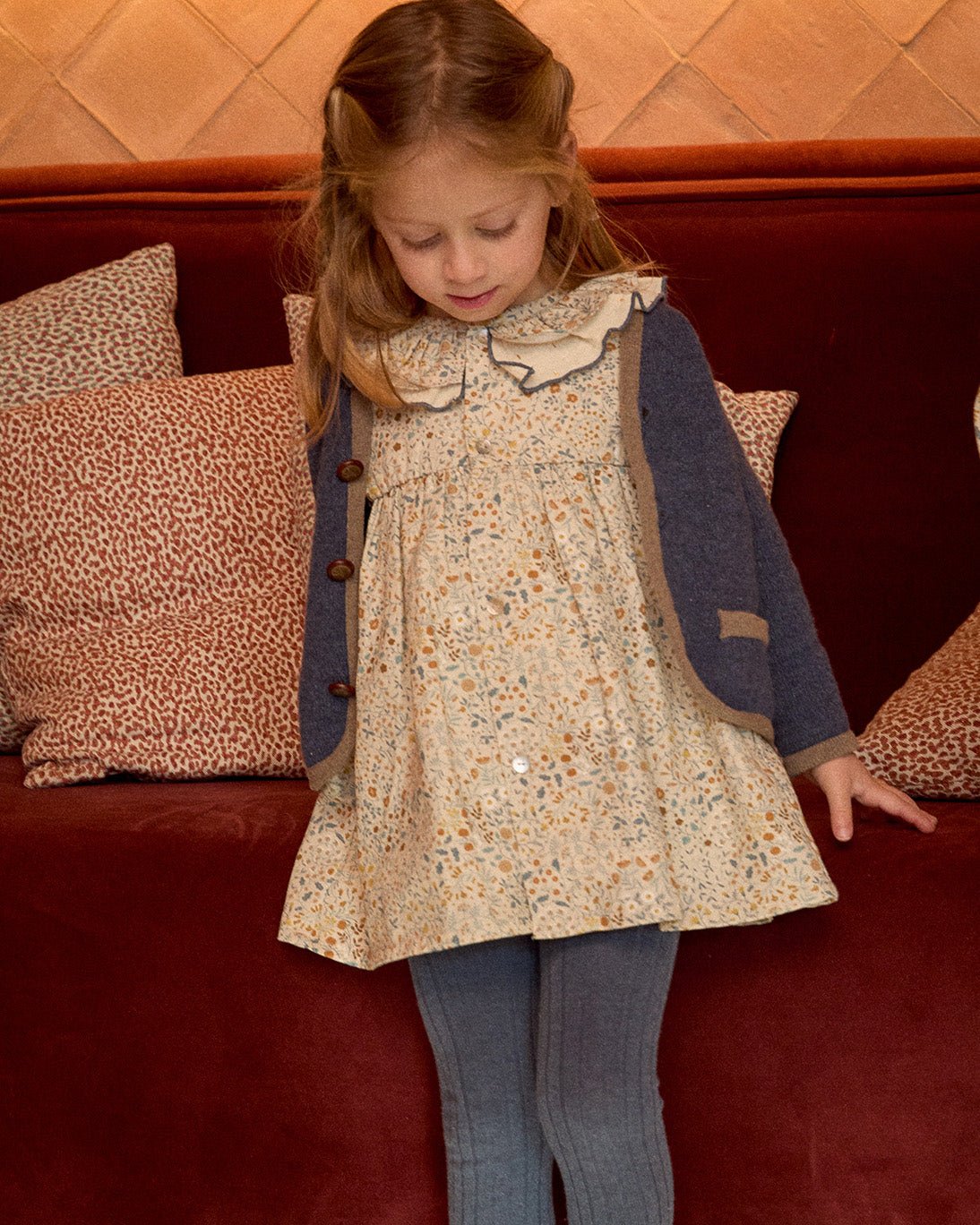 Young girl in a floral dress and blue cardigan standing on a red carpet with patterned pillows in the background.