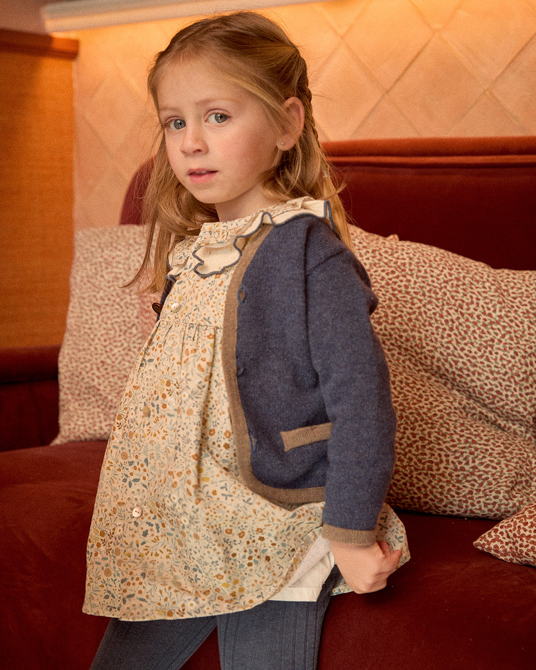 Young girl wearing a patterned dress and blue cardigan standing in front of a red couch with pillows.