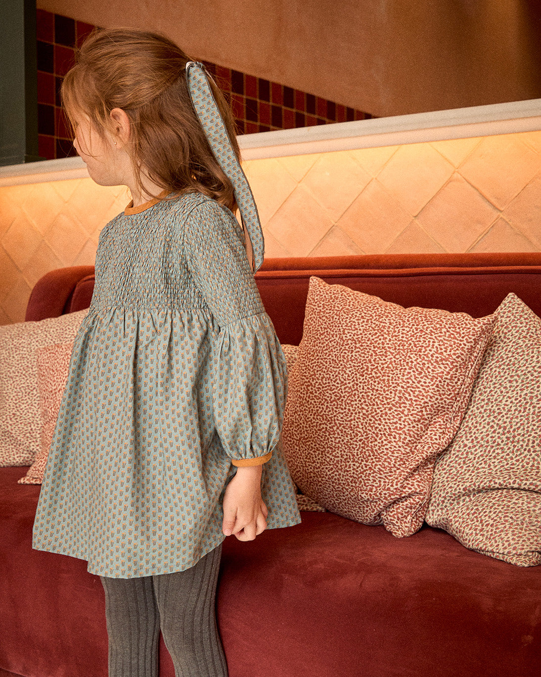 Child in a patterned dress standing next to a red couch with pillows.