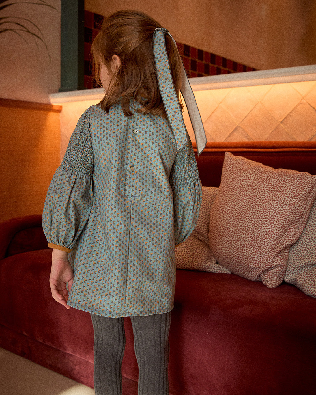 Person wearing a patterned coat standing in front of a red couch with pillows.