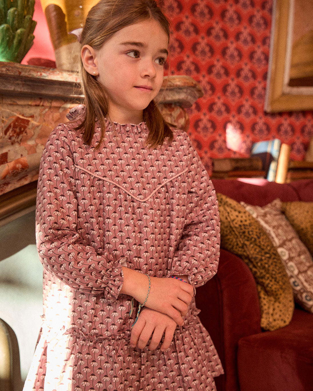 Young girl in a pink textured dress standing in a room with patterned wallpaper and furniture.