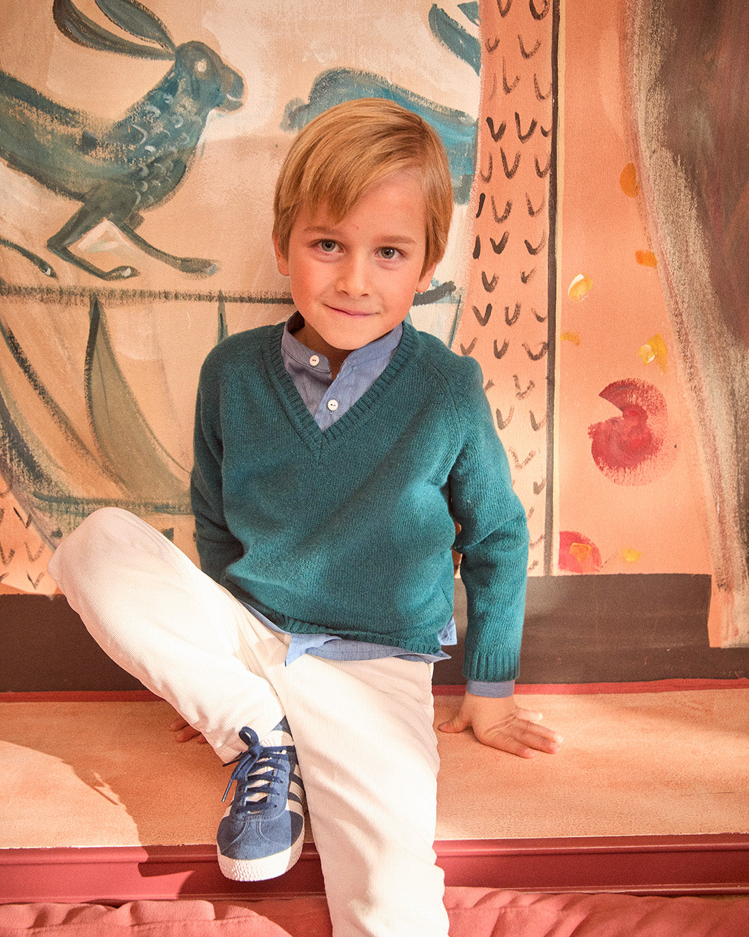 Young boy in a teal sweater and white pants sitting on a colorful rug.