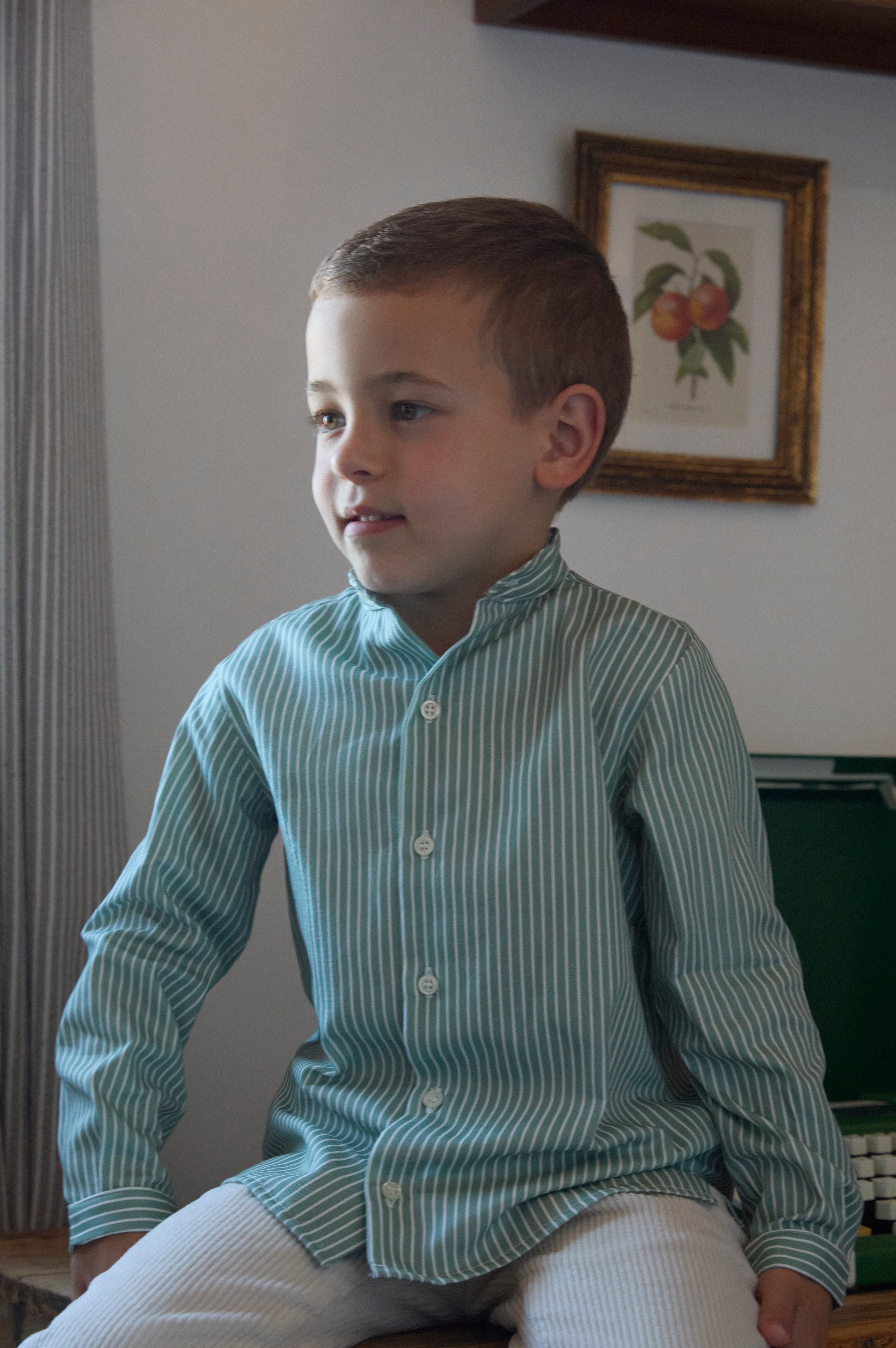 Young boy sitting on a wooden bench wearing a green striped shirt in a room with framed pictures on the wall.