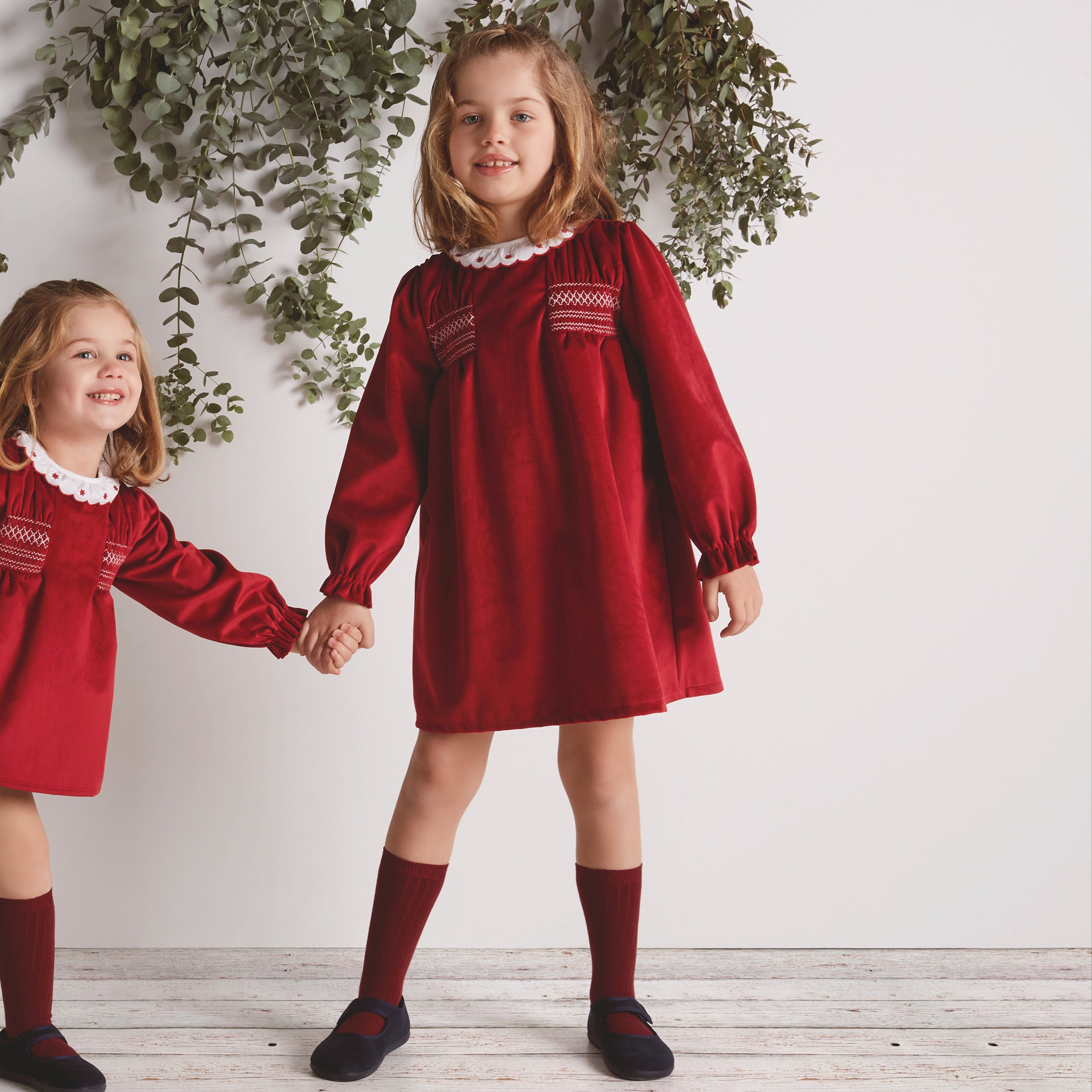 Two young girls in red dresses standing against a white wall with greenery.