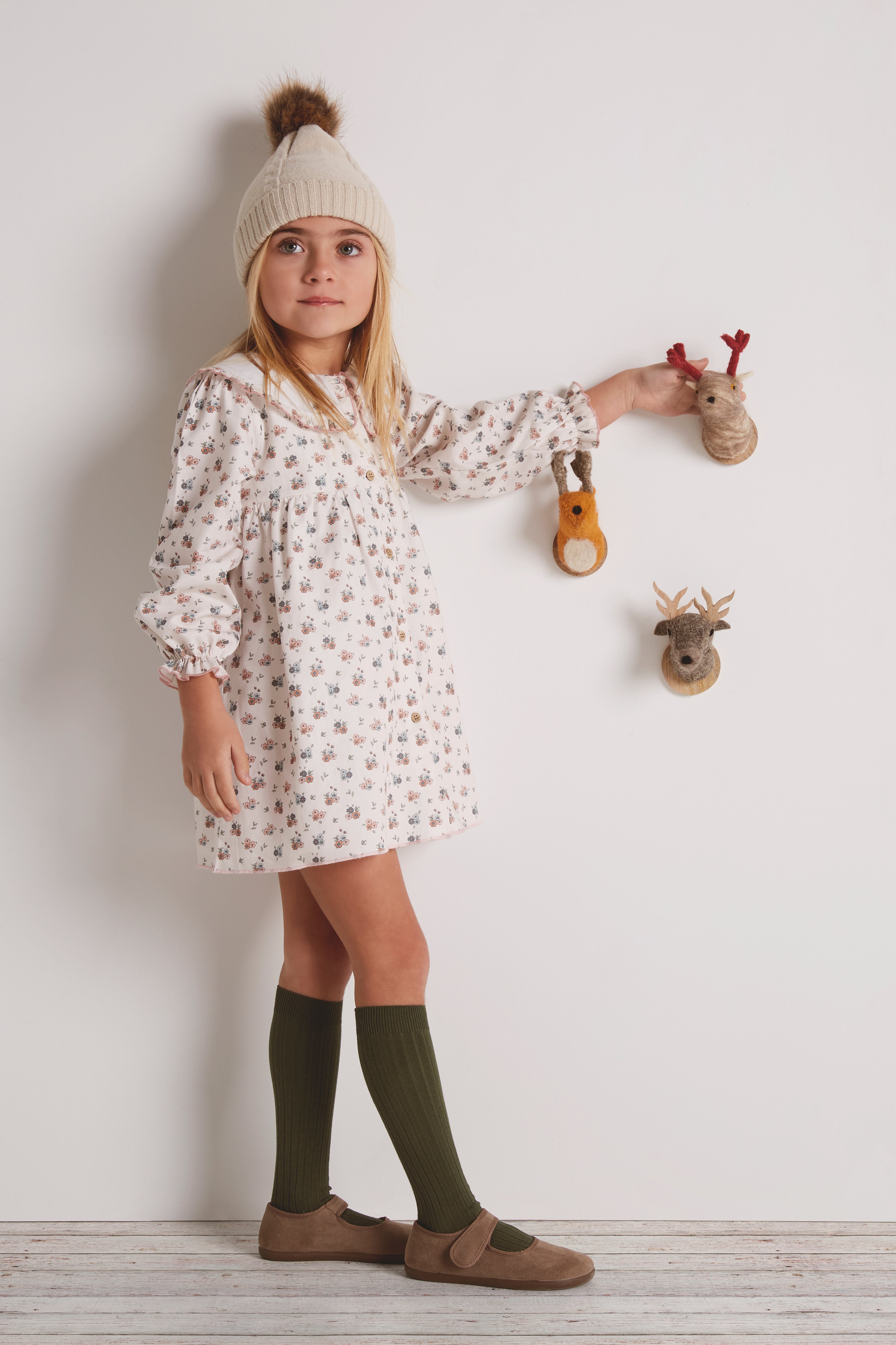 Young girl in a floral dress with green socks and brown shoes standing against a white wall.