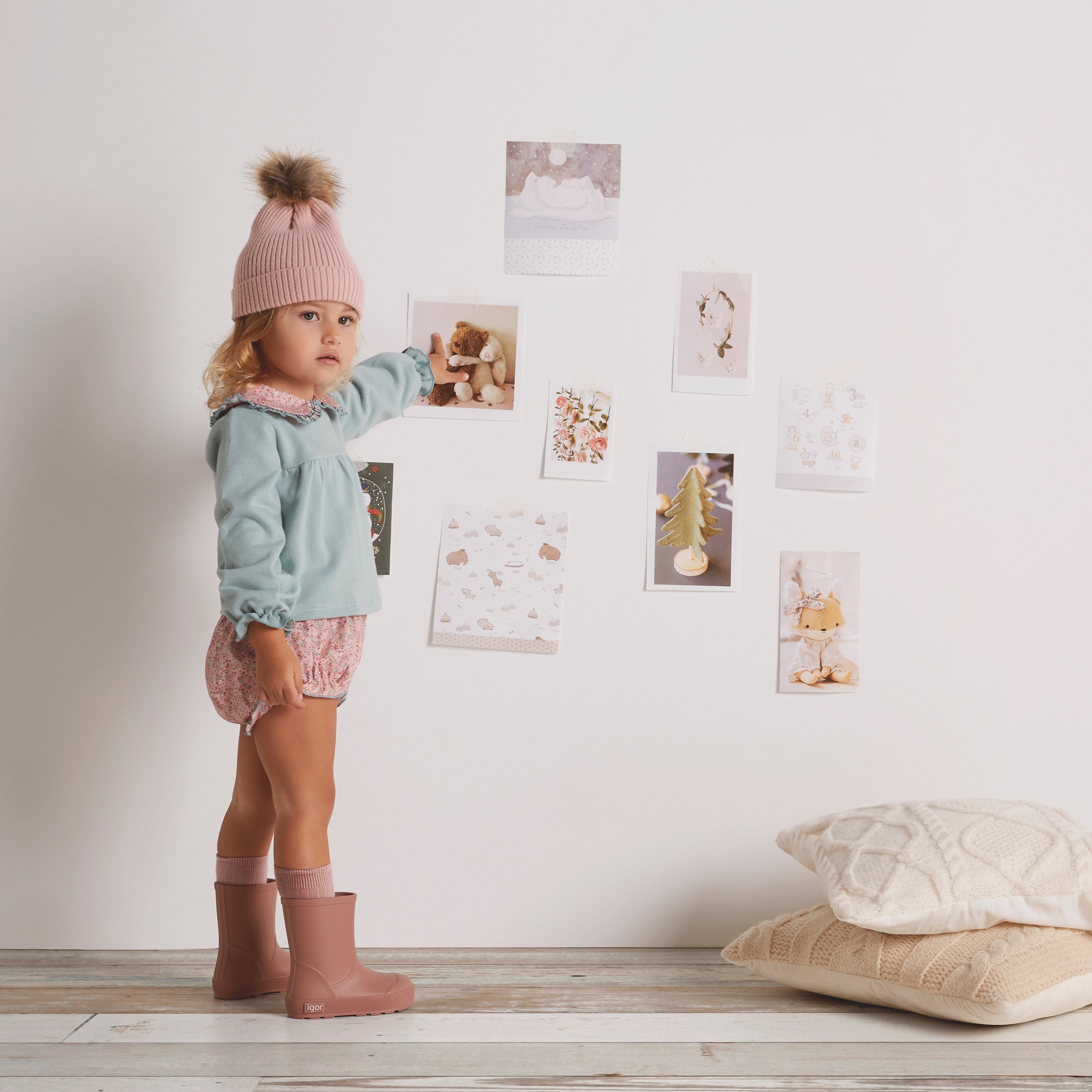 Child standing in a room with a white wall, wearing a light blue sweater and pink boots.
