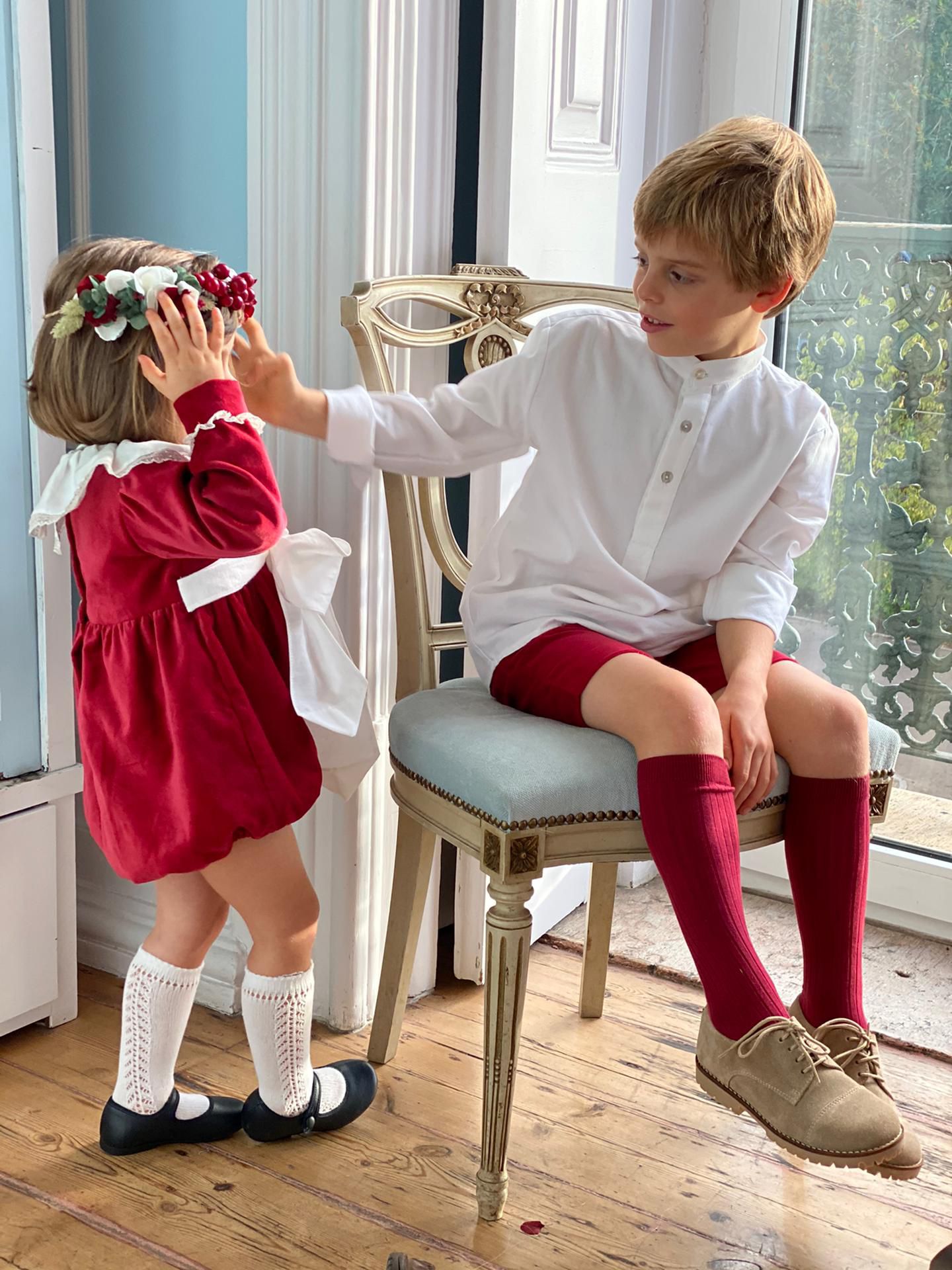Two children, a girl in a red dress and a boy in a white shirt and red knee-high socks, interacting indoors.