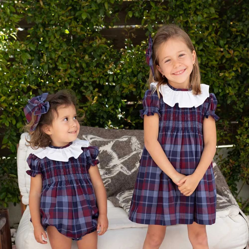 Two young girls in matching plaid dresses with white collars standing outdoors.