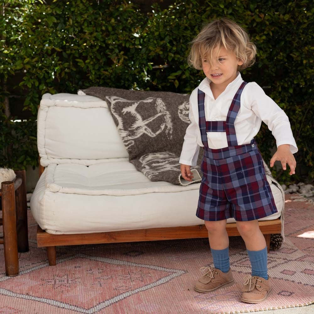 Child wearing a white shirt, plaid overalls, and blue socks standing in front of a white cushioned chair outdoors.