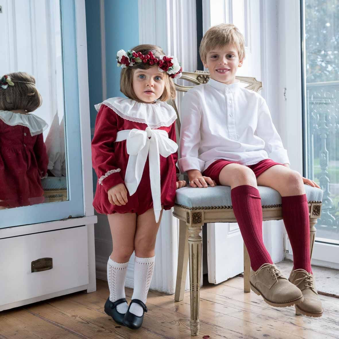 Two children in matching outfits sitting on a chair in a room with a mirror.